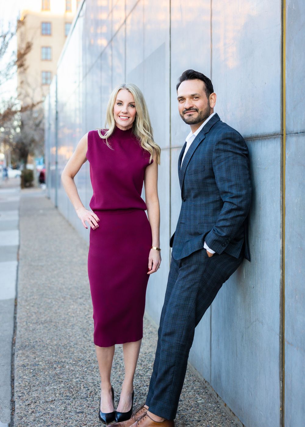A couple poses against a modern building exterior with the person wearing a burgundy dress and navy suit.