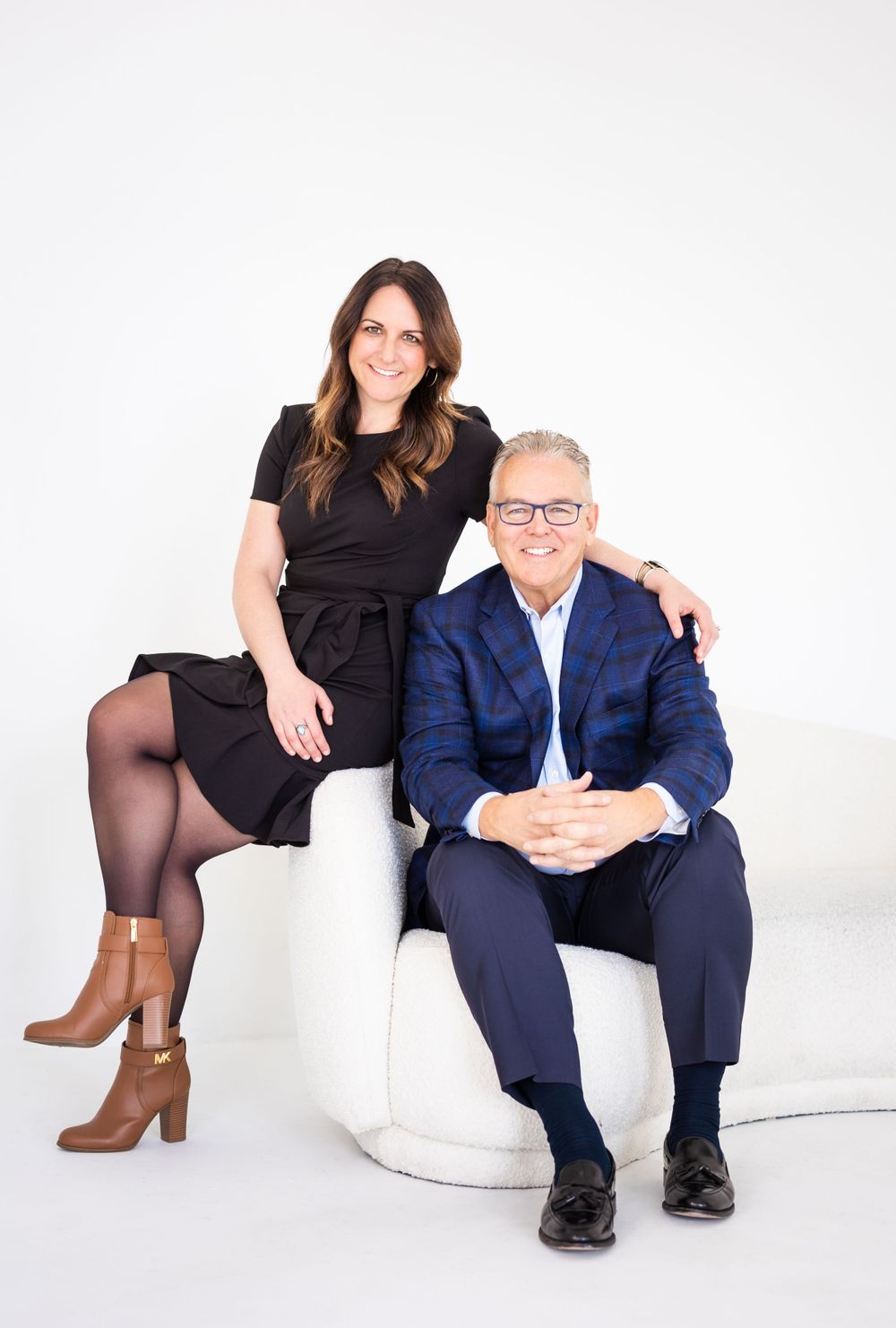 Studio photo of two people seated on white furniture in professional business attire.