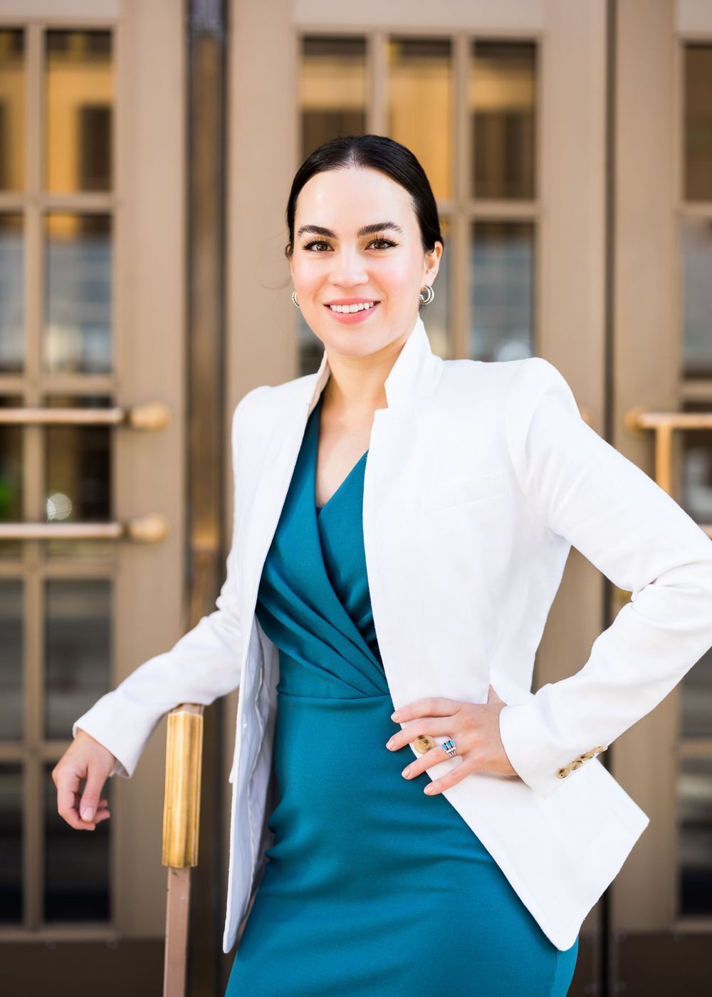 Professional woman in white blazer and teal dress poses confidently against office building exterior.
