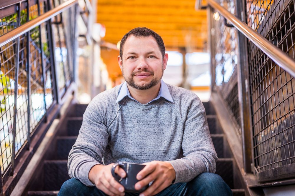A person in a gray sweater sitting on steps holding a cup and smiling warmly at the camera.