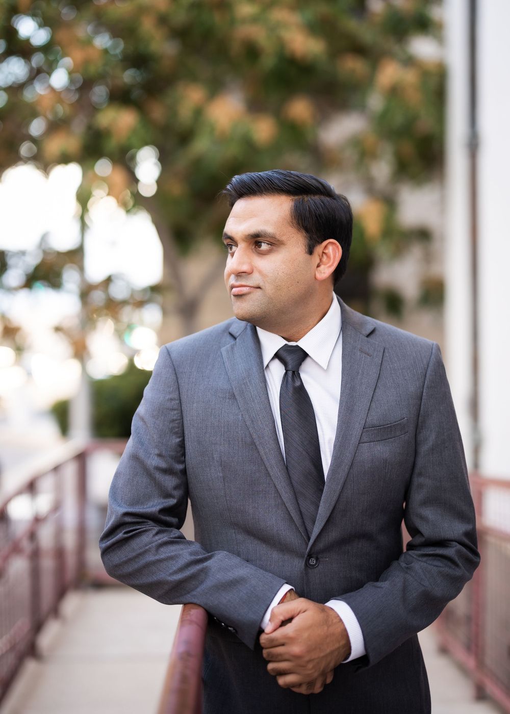 Professional headshot of person in grey suit on outdoor walkway.