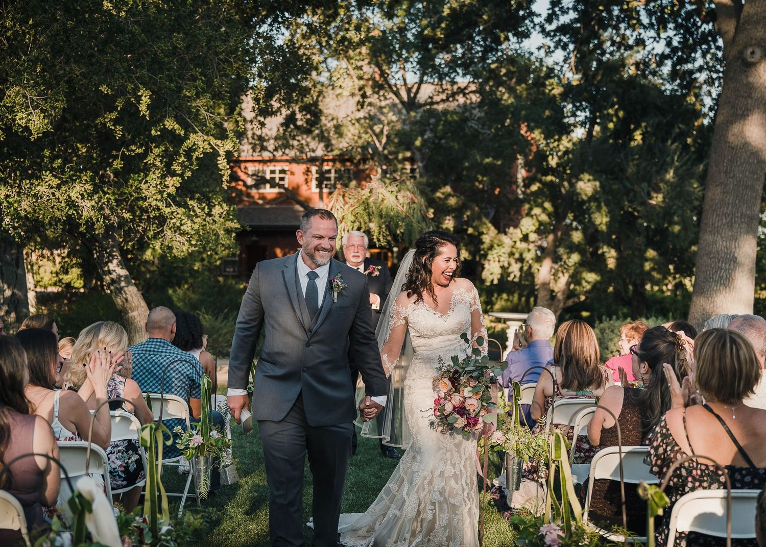 A couple walks down the aisle after their outdoor ceremony while guests seated on white chairs look on.