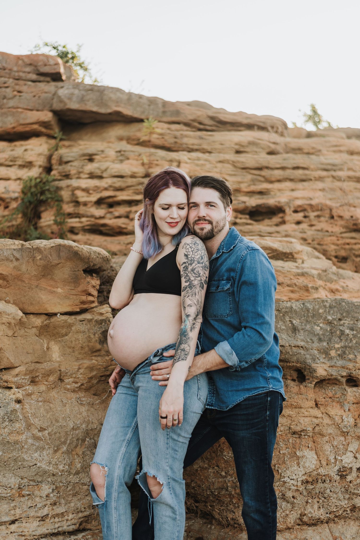 A maternity photoshoot against red rock formations shows a couple in casual denim embracing the pregnancy moment.