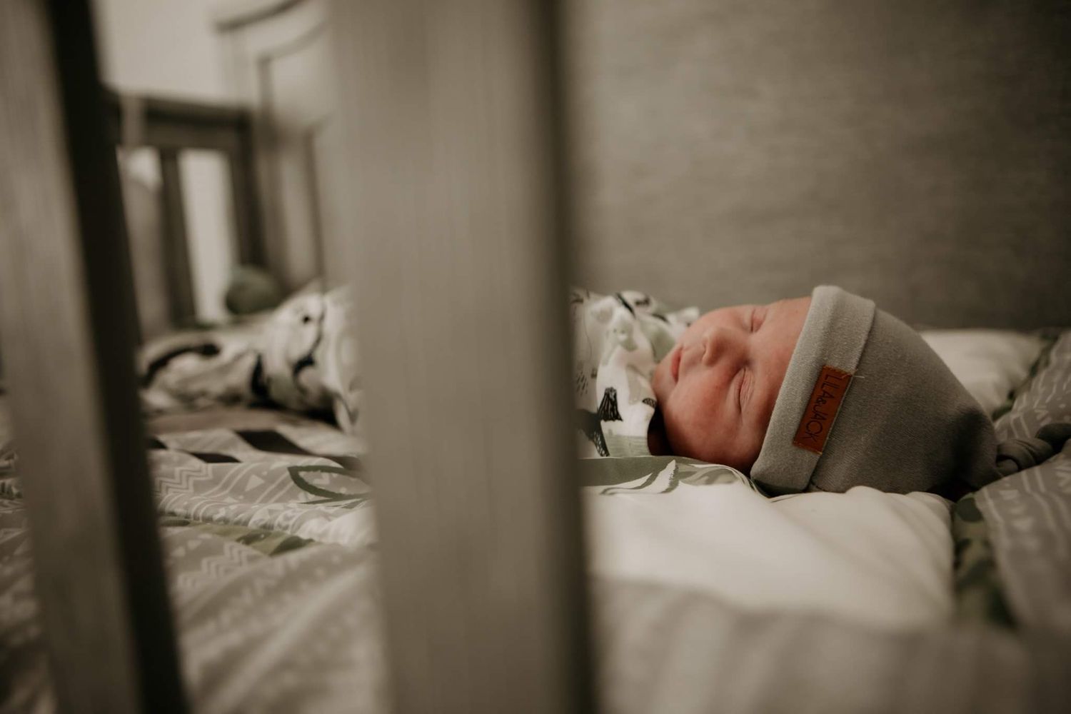 A sleeping newborn in a gray hat viewed through crib slats in black and white.