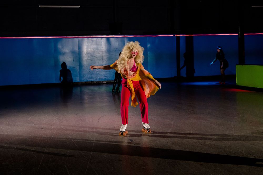 Roller skater in bright orange and red outfit performs dramatic move on dark rink.