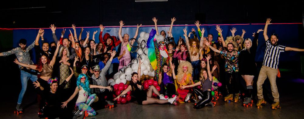 Large group of roller derby participants posing together with raised arms in celebration on a dark rink.