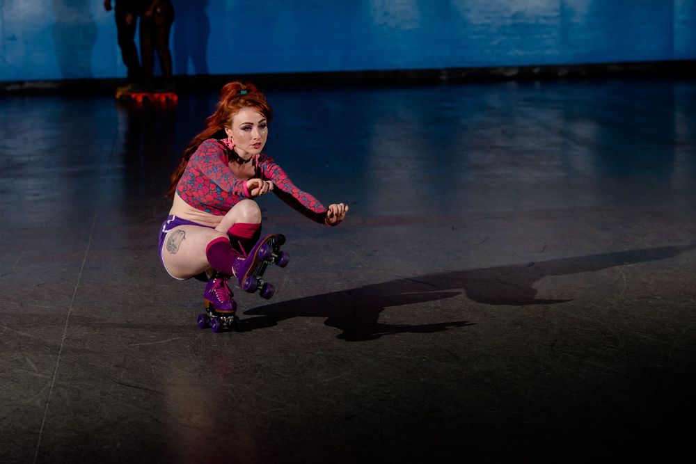 A skater in purple clothing crouches low while performing on a dark roller rink.