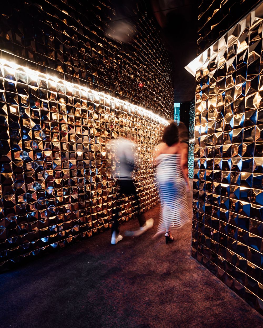 Two blurry figures walk through a hallway with metallic textured walls illuminated by dramatic lighting.