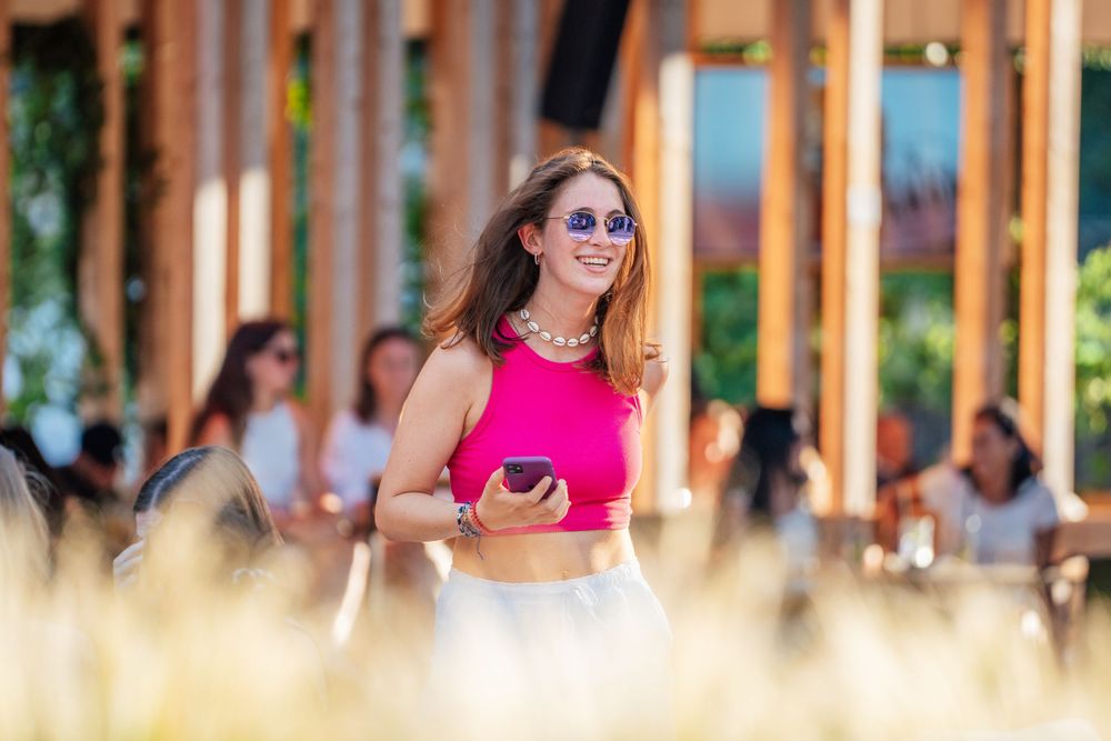 Person in bright pink crop top and sunglasses stands outdoors with blurred foreground grass and wooden columns.
