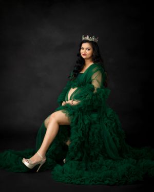 Glamorous maternity portrait with emerald gown, crown, and silver heels against dark backdrop.
