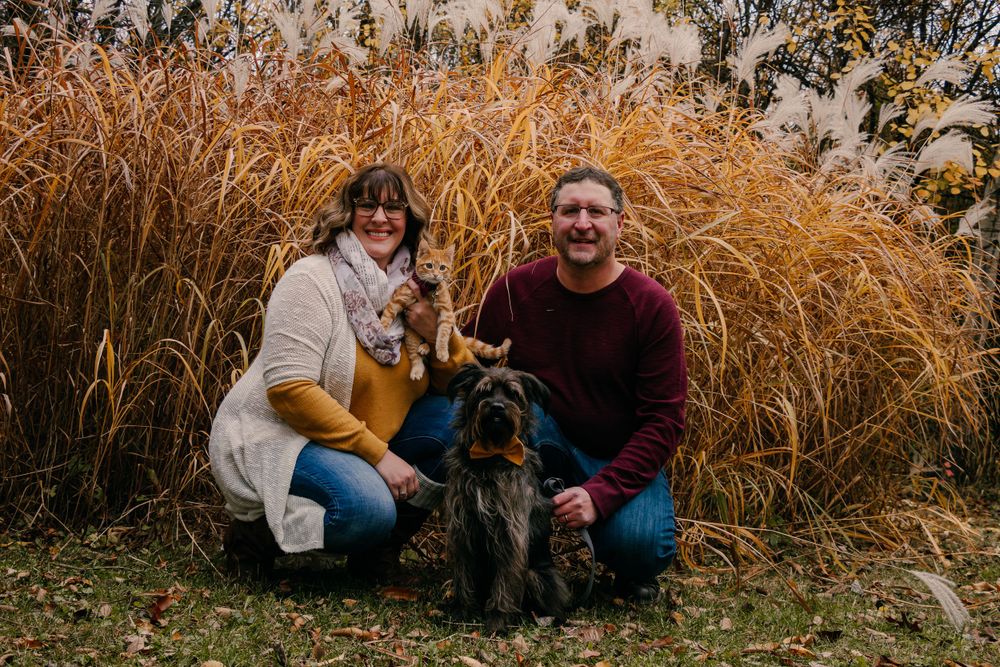 Couple sitting with their black dog in front of tall autumn grasses.
