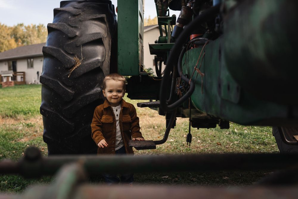 Small toddler in brown coat playing near large tractor tire in rural farm setting.