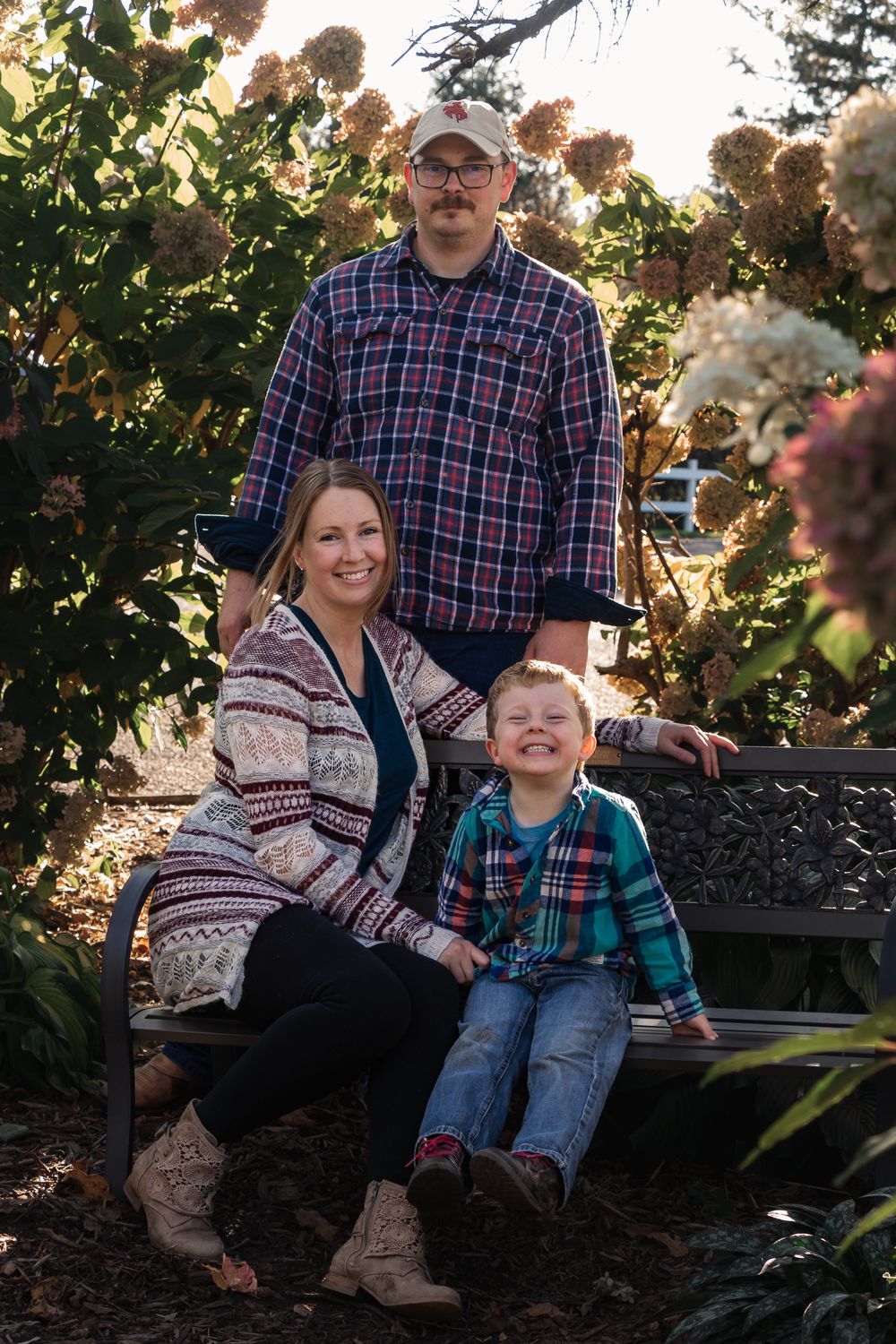 Family poses together on a garden bench surrounded by fall foliage and flowering bushes.