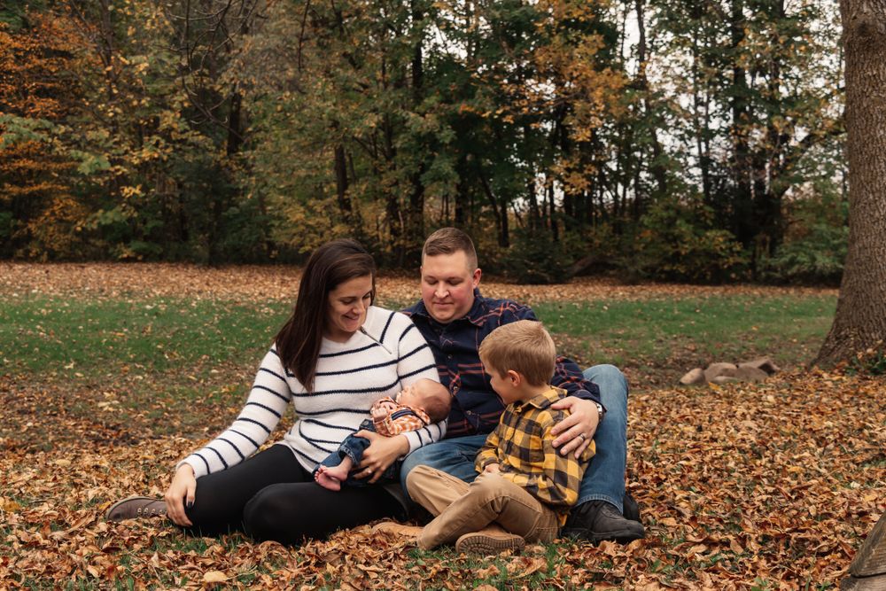 Family relaxes together on fallen autumn leaves in a wooded park setting.