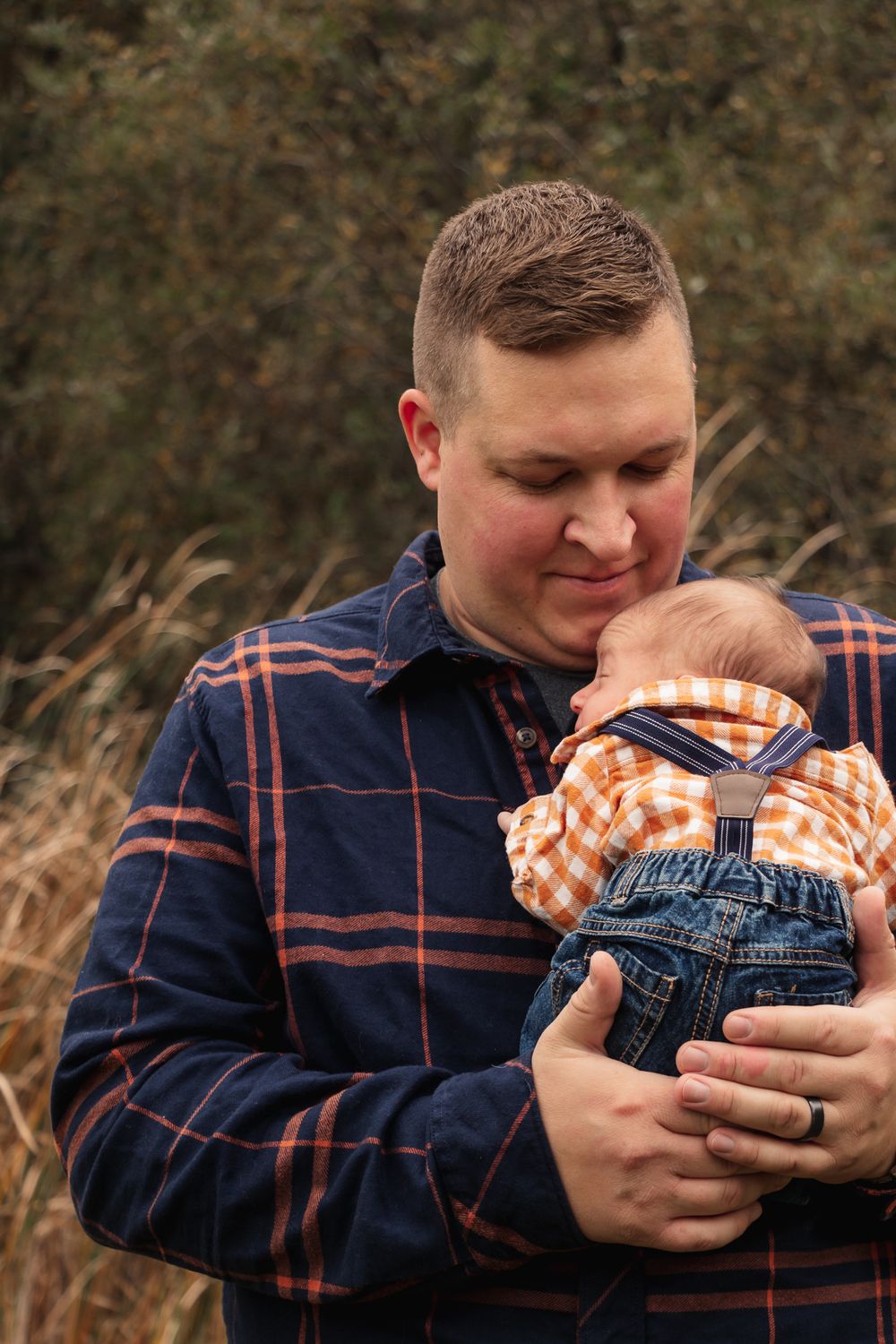 Parent tenderly holds sleeping infant wearing orange plaid shirt against plaid jacket.