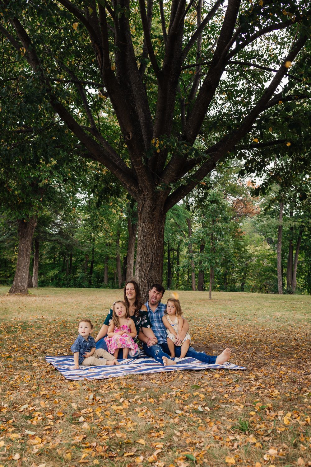 Family group lounging on blankets under large autumn trees with fallen leaves scattered around.