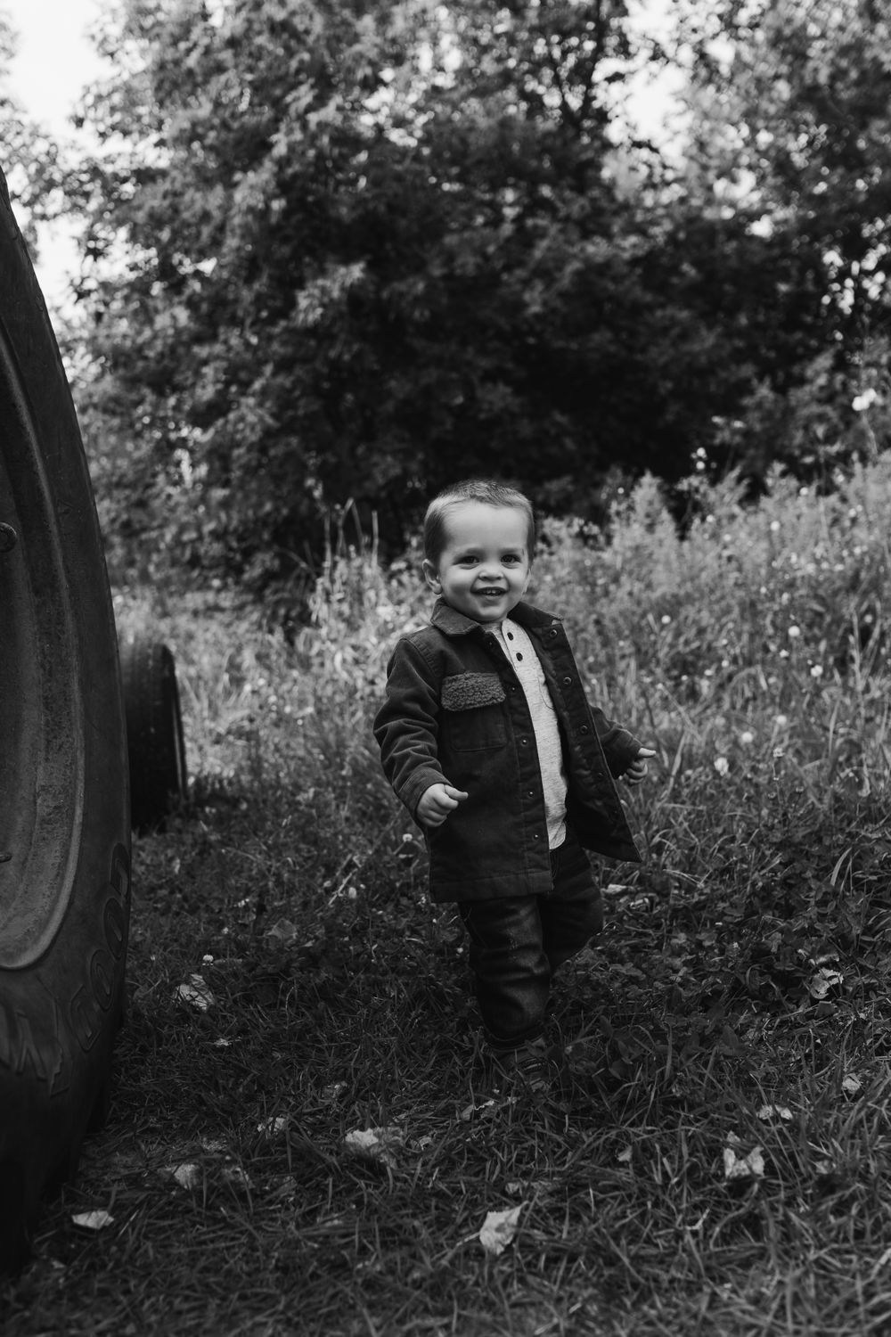 Black and white photo of toddler standing by large tractor wheel in grass.