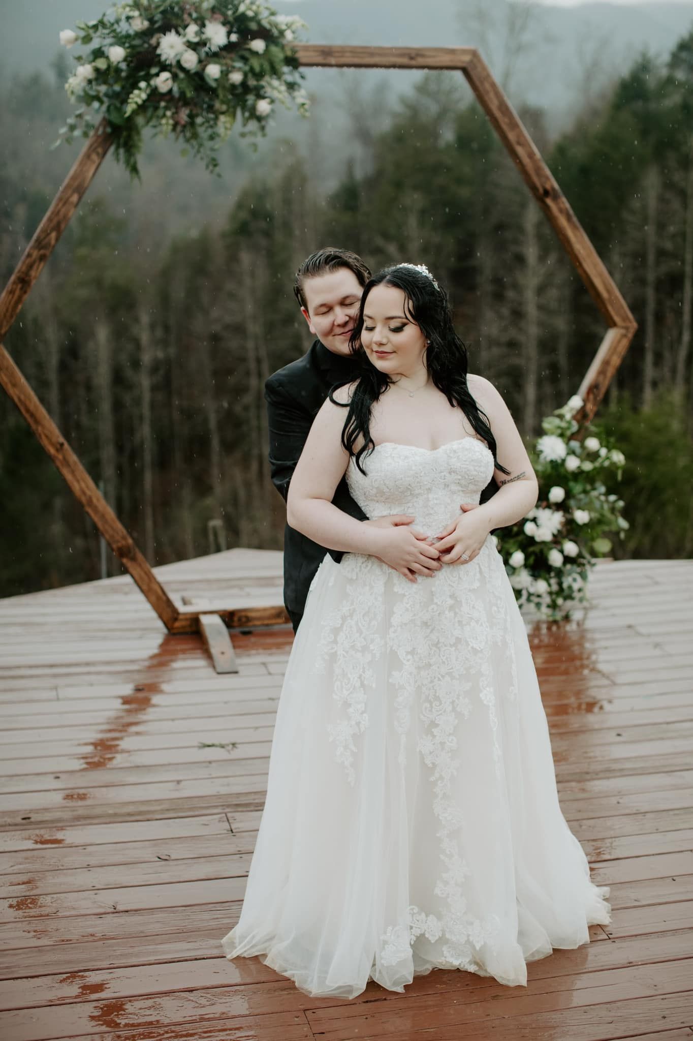 Wedding couple embracing on outdoor deck with mountain scenery and wooden arch.