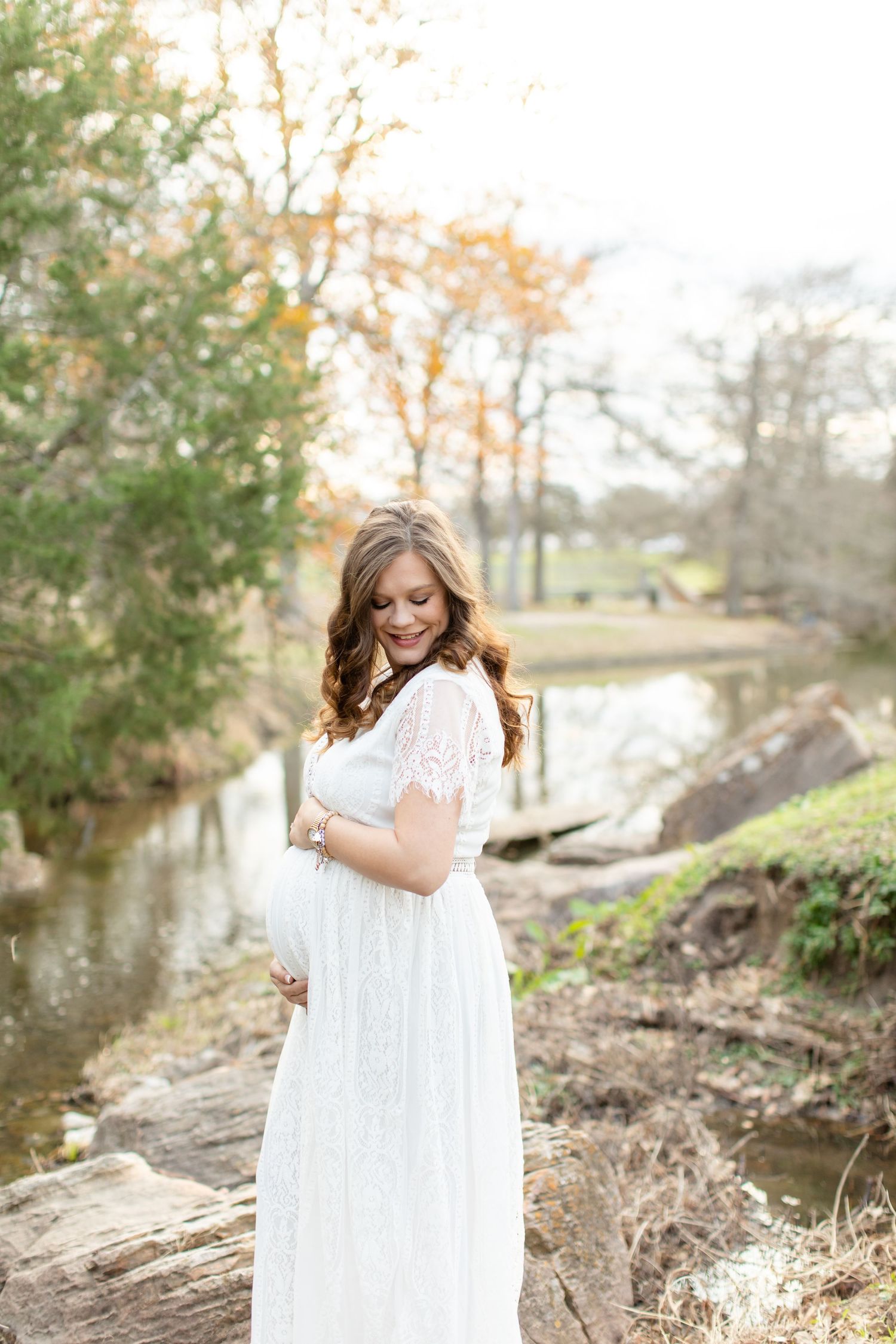 An expectant mother in white lace dress poses beside a peaceful stream.