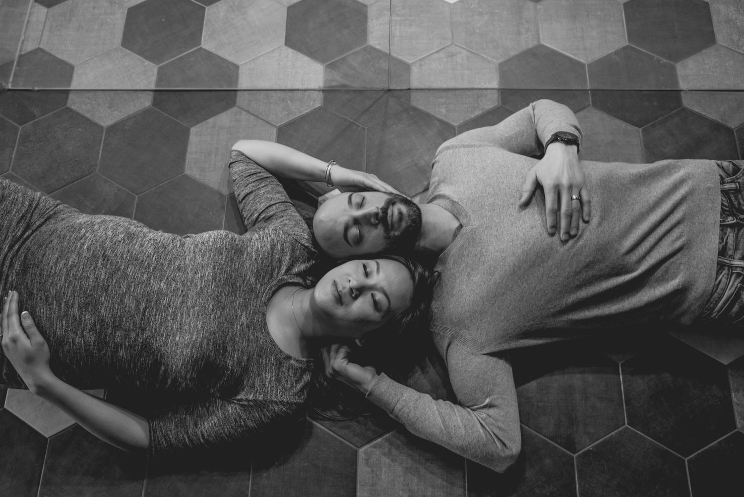 A black and white photo showing two people lying on a hexagonal tiled floor in casual poses.