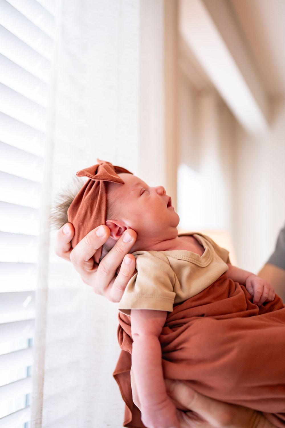 Newborn baby wearing tan outfit and rust-colored headband being held near window.