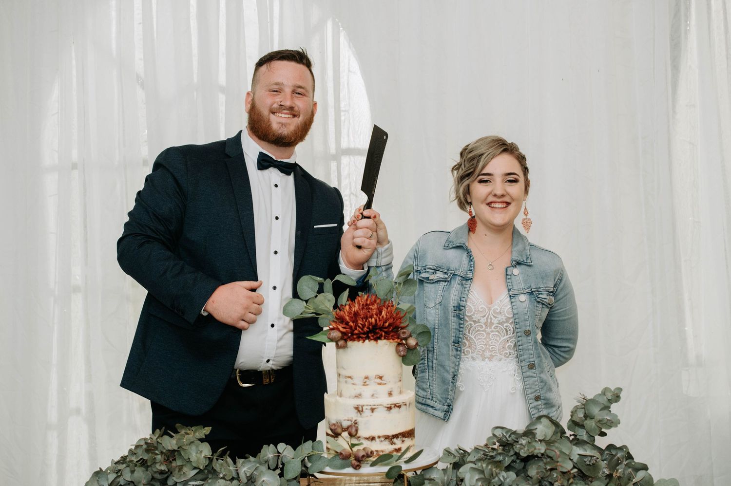 A newlywed couple poses with their rustic wedding cake decorated with red flowers and greenery.