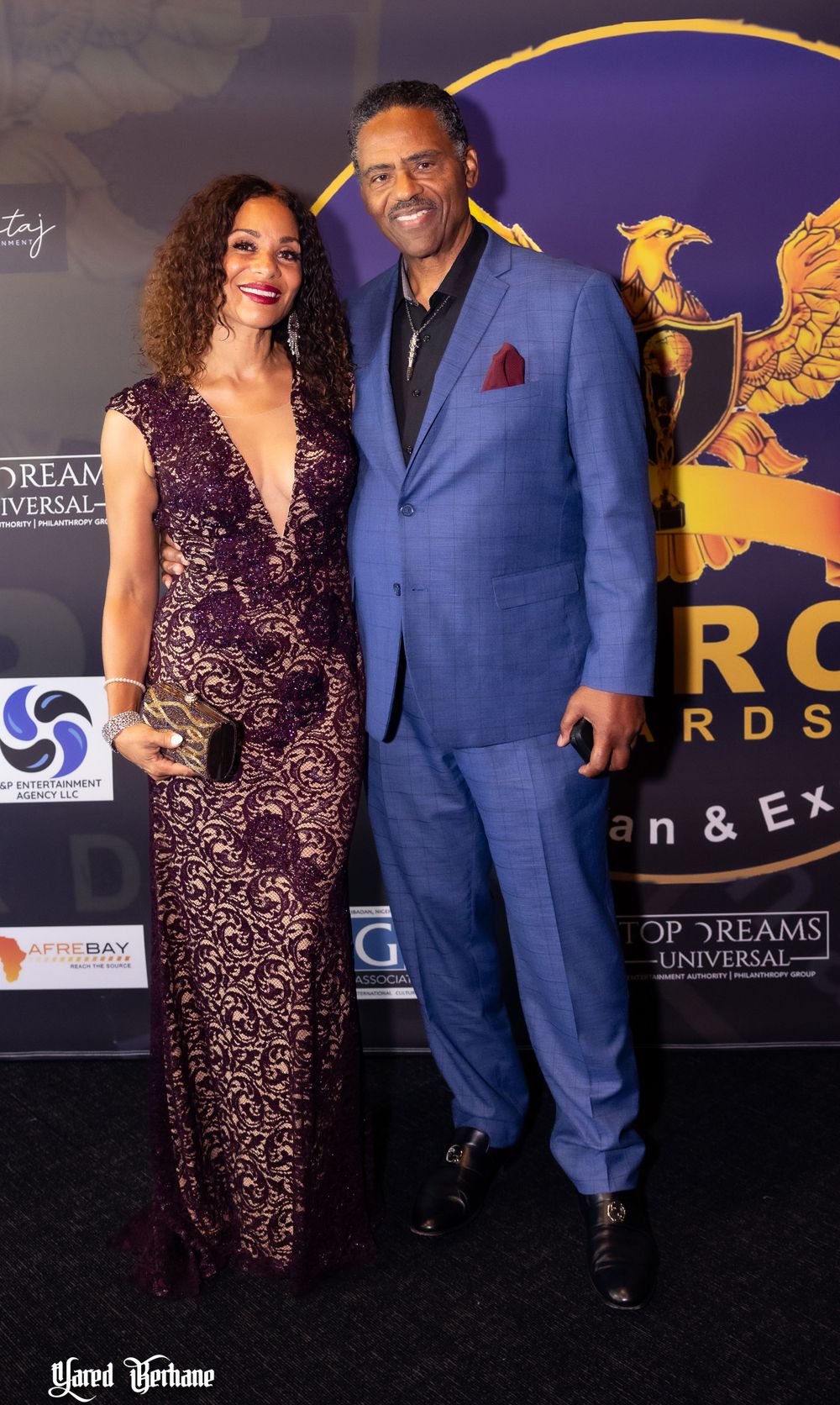 Elegant couple in purple gown and blue suit pose at awards ceremony against branded backdrop.