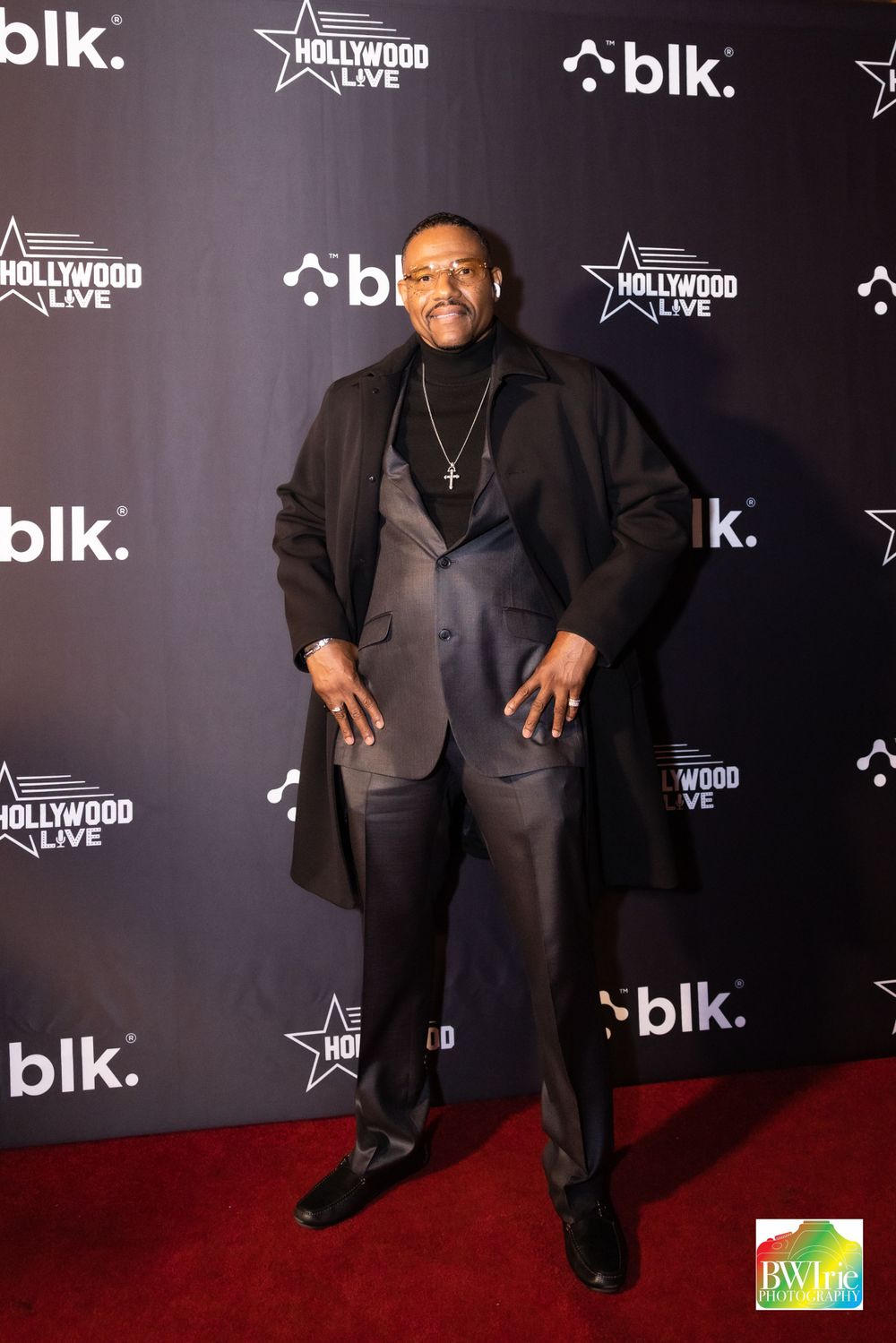A guest in a black suit and overcoat poses on the red carpet at a BLK entertainment event with Hollywood Live branding.