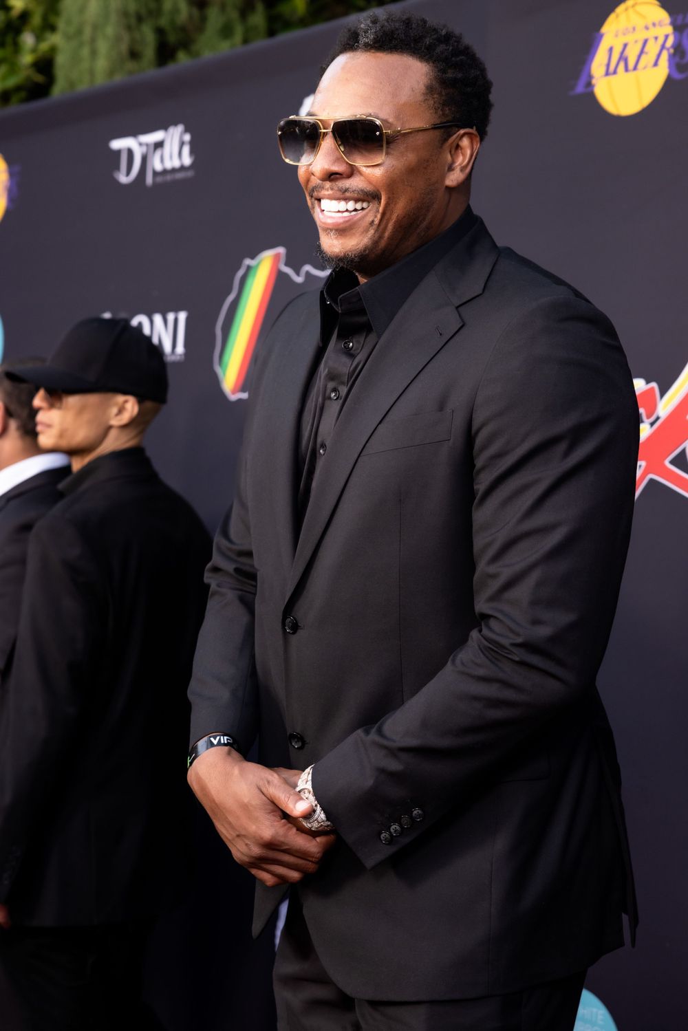 A performer in a black suit and sunglasses smiles while posing on a red carpet at a Hollywood event.