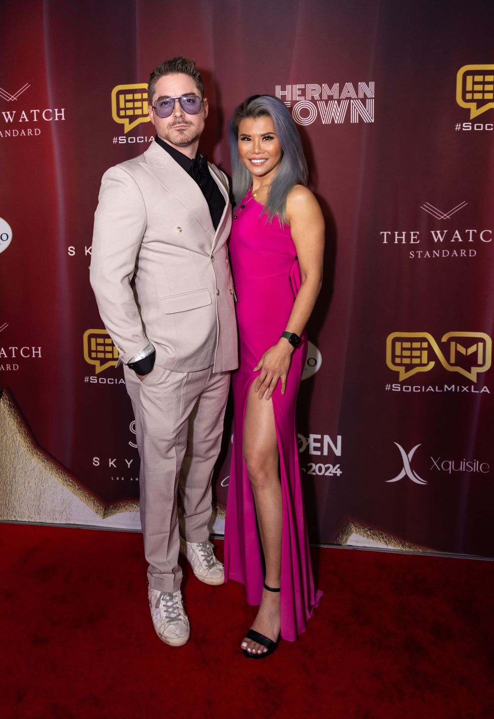 Couple poses at red carpet event with man in beige suit and woman in pink high-slit gown.