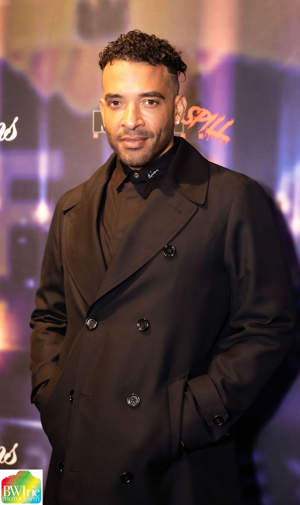 Man in dark double-breasted coat poses at entertainment industry event.