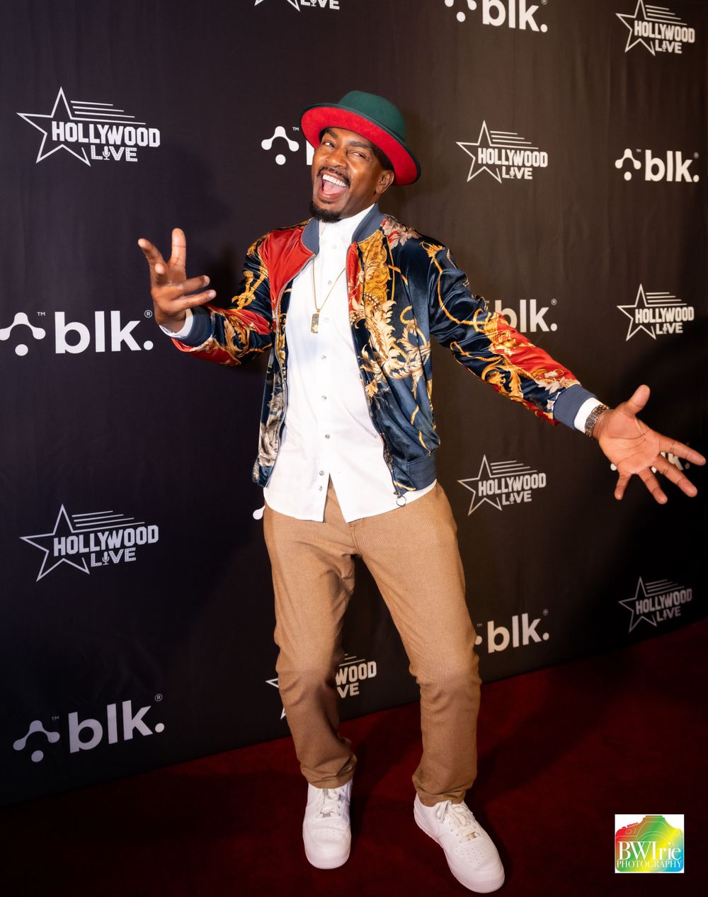 Performer in vibrant jacket and red hat strikes animated pose on red carpet at BLK event.