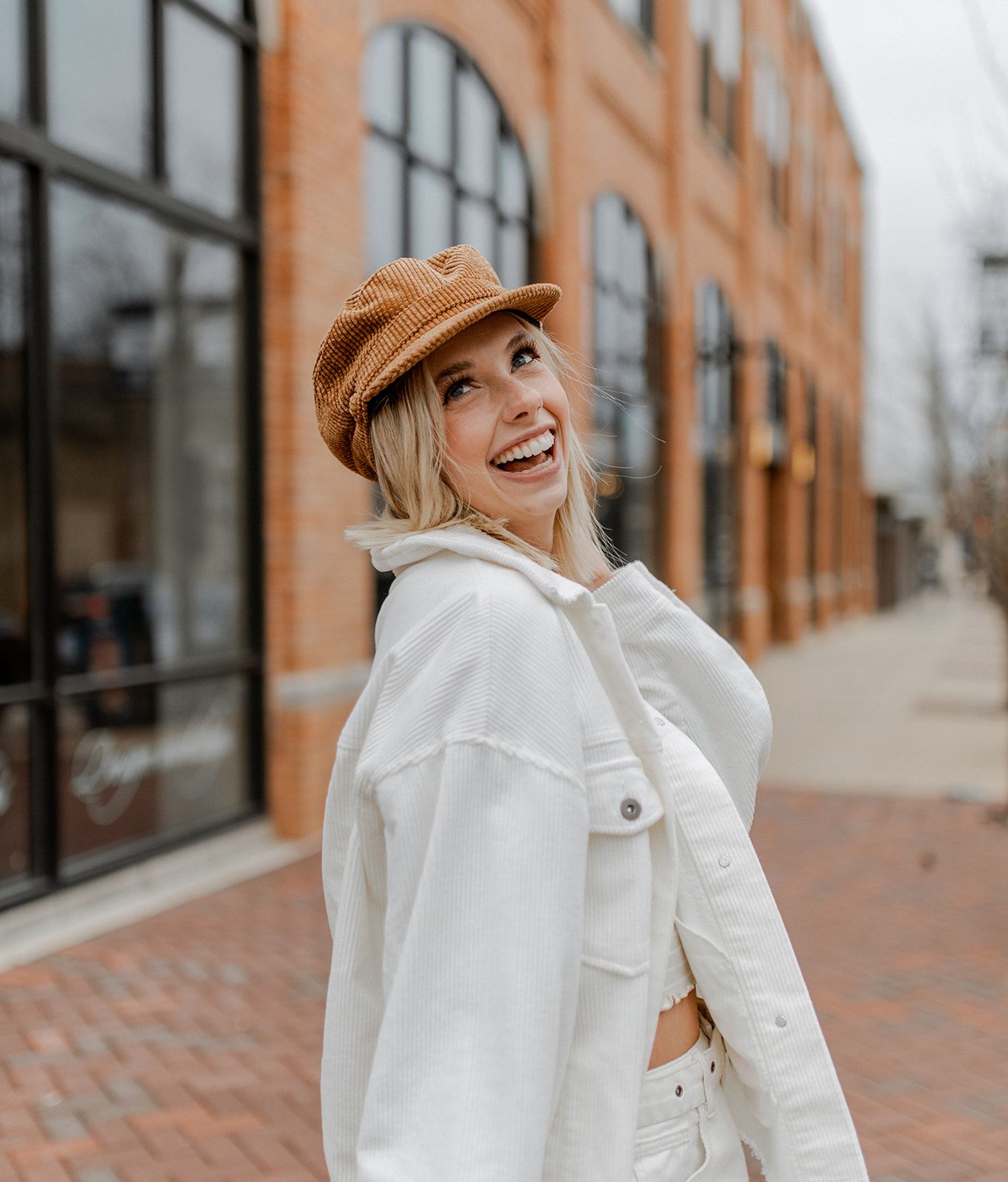 A joyful pose in white outerwear and tan newsboy cap against an urban brick building backdrop.