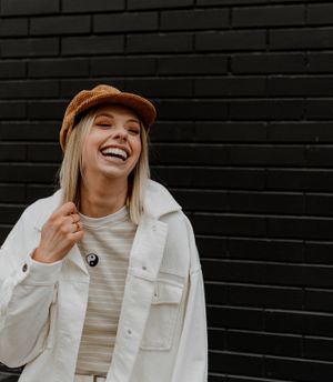 Casual street style portrait against black brick wall wearing white jacket and tan cap.