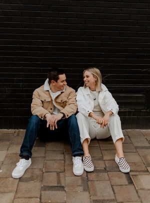 Couple in coordinated outfits sitting and laughing against black brick wall.