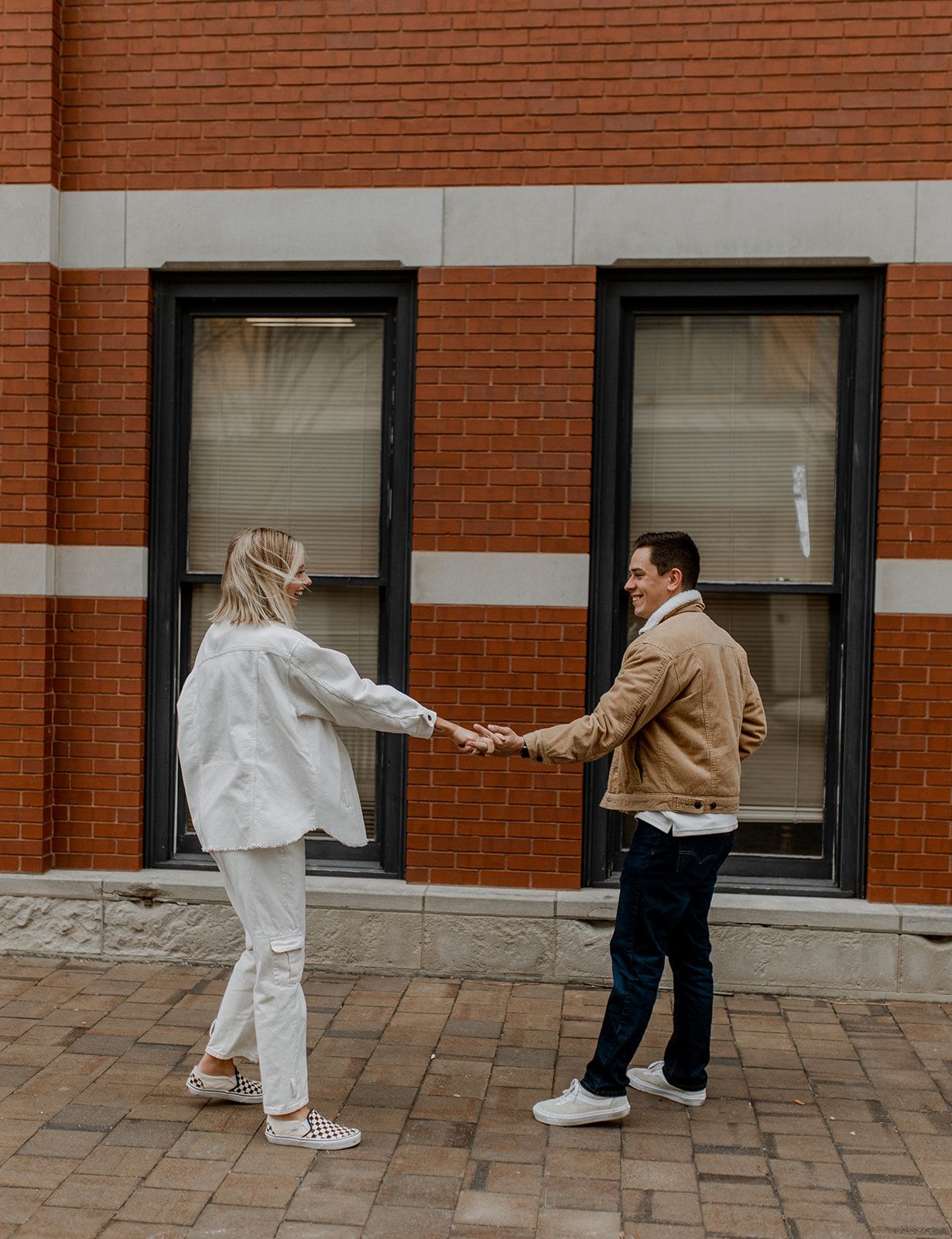 Couple in white and tan outerwear holding hands while walking on brick pavement by building.