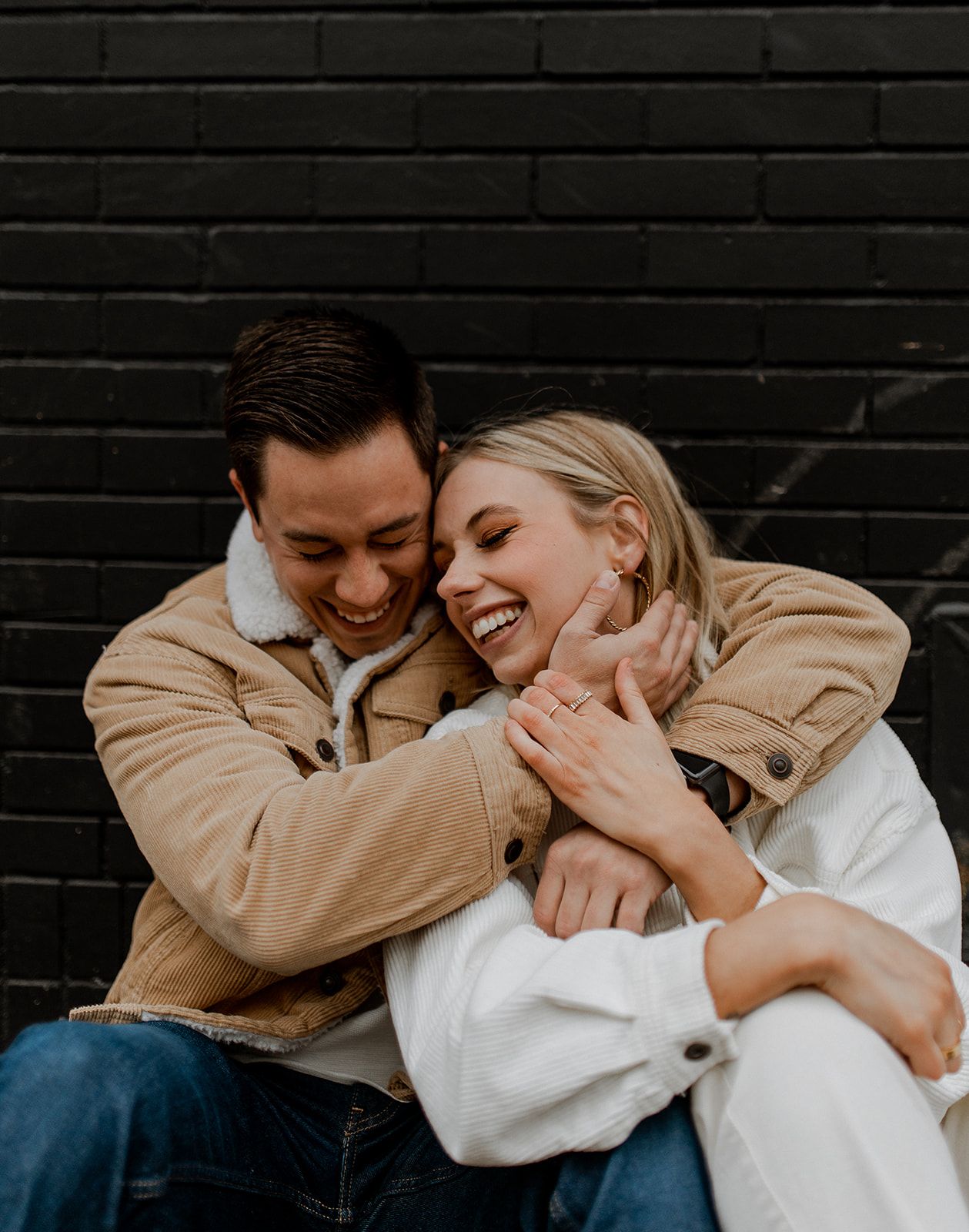 Romantic couple sharing an intimate embrace against a dark brick wall background, laughing together joyfully.