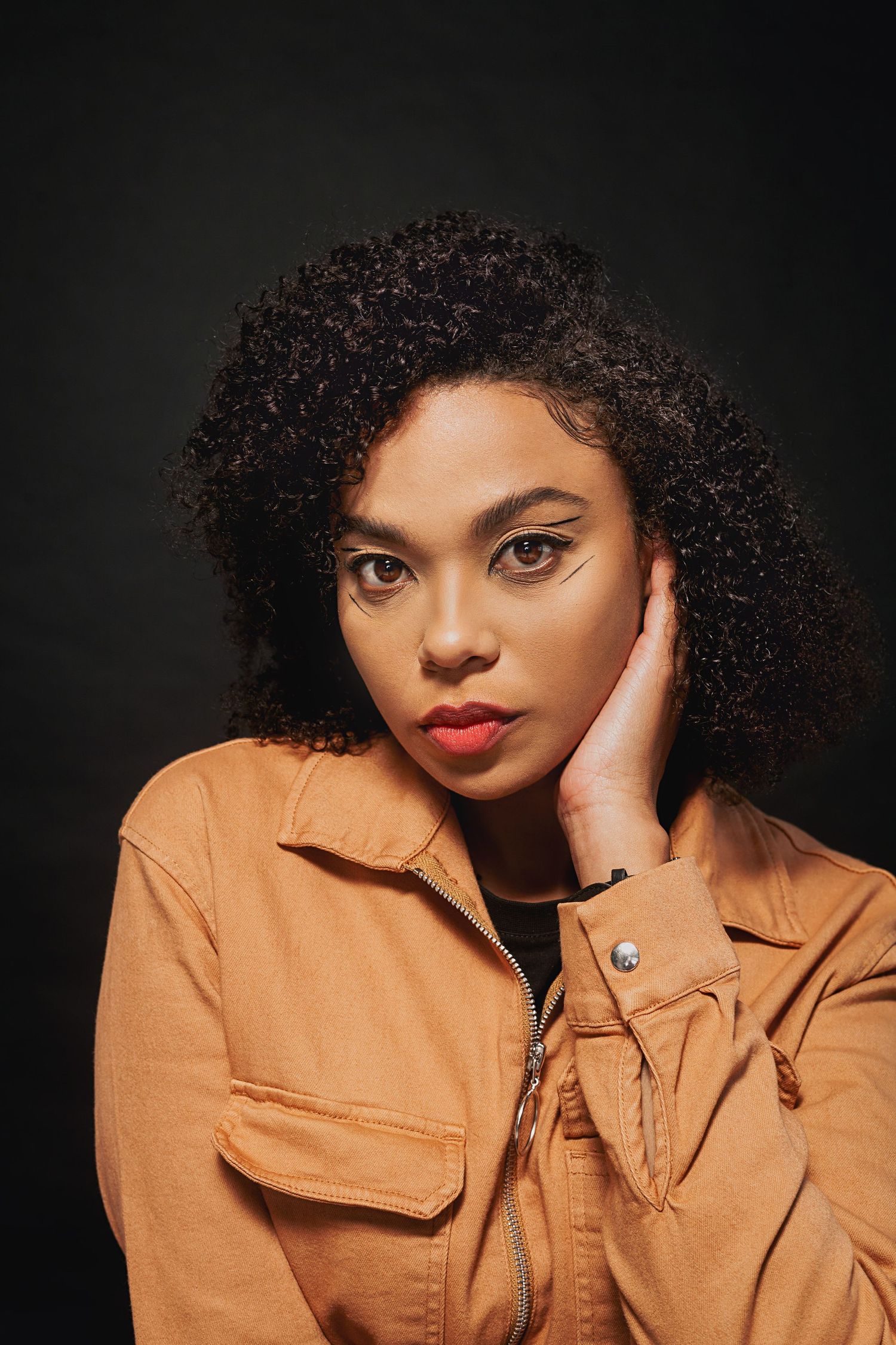 Beauty portrait featuring natural curly hair and caramel utility jacket against dark backdrop.