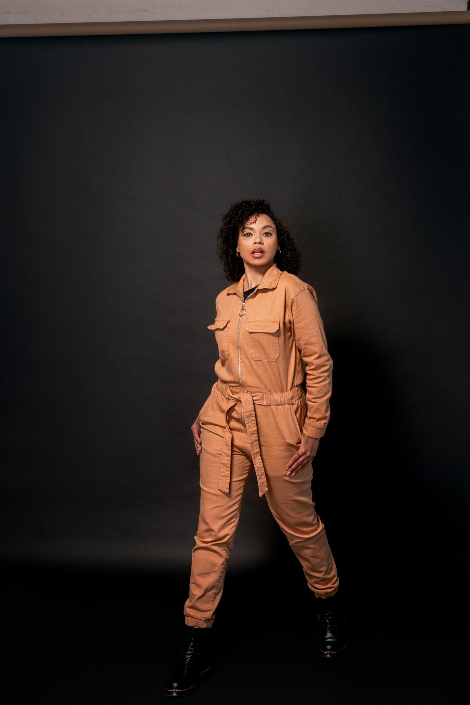 Fashion model poses in tan utility jumpsuit against dark background.