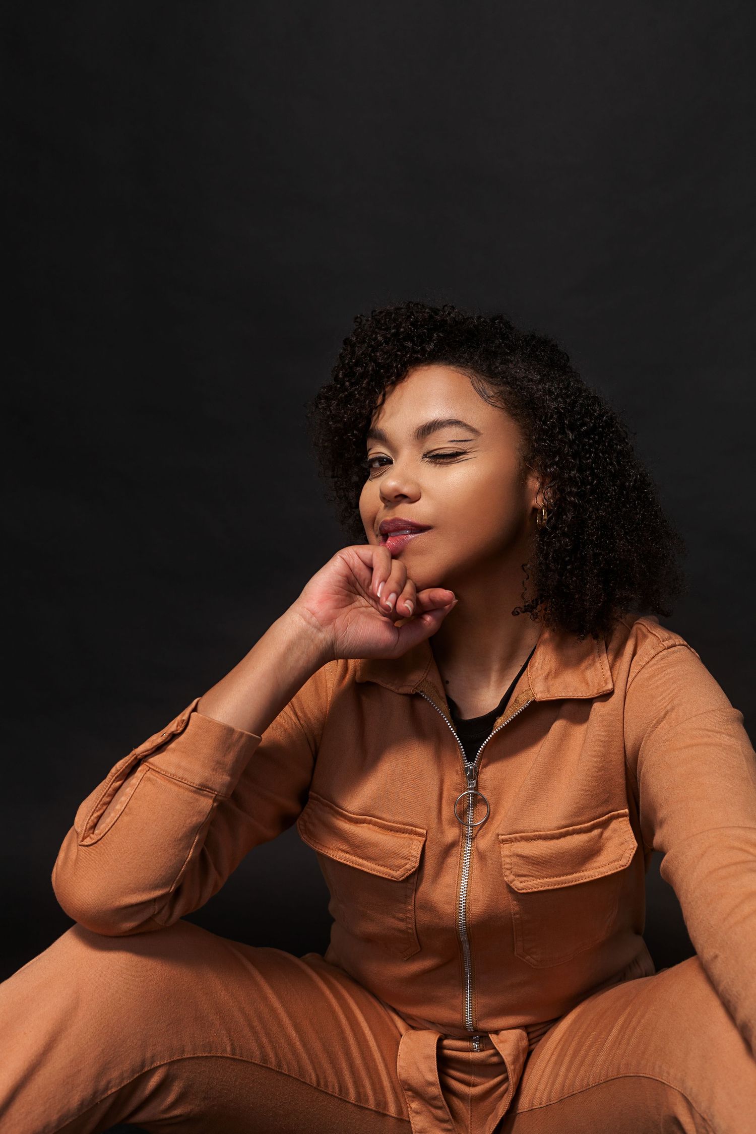 Dramatic portrait series in caramel-colored jumpsuit against black backdrop.