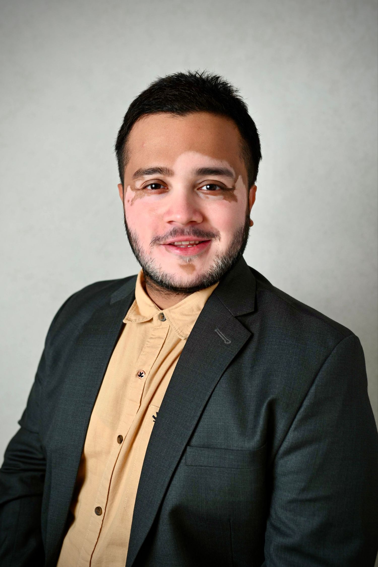 Business portrait of person in tan shirt and dark blazer with facial hair.