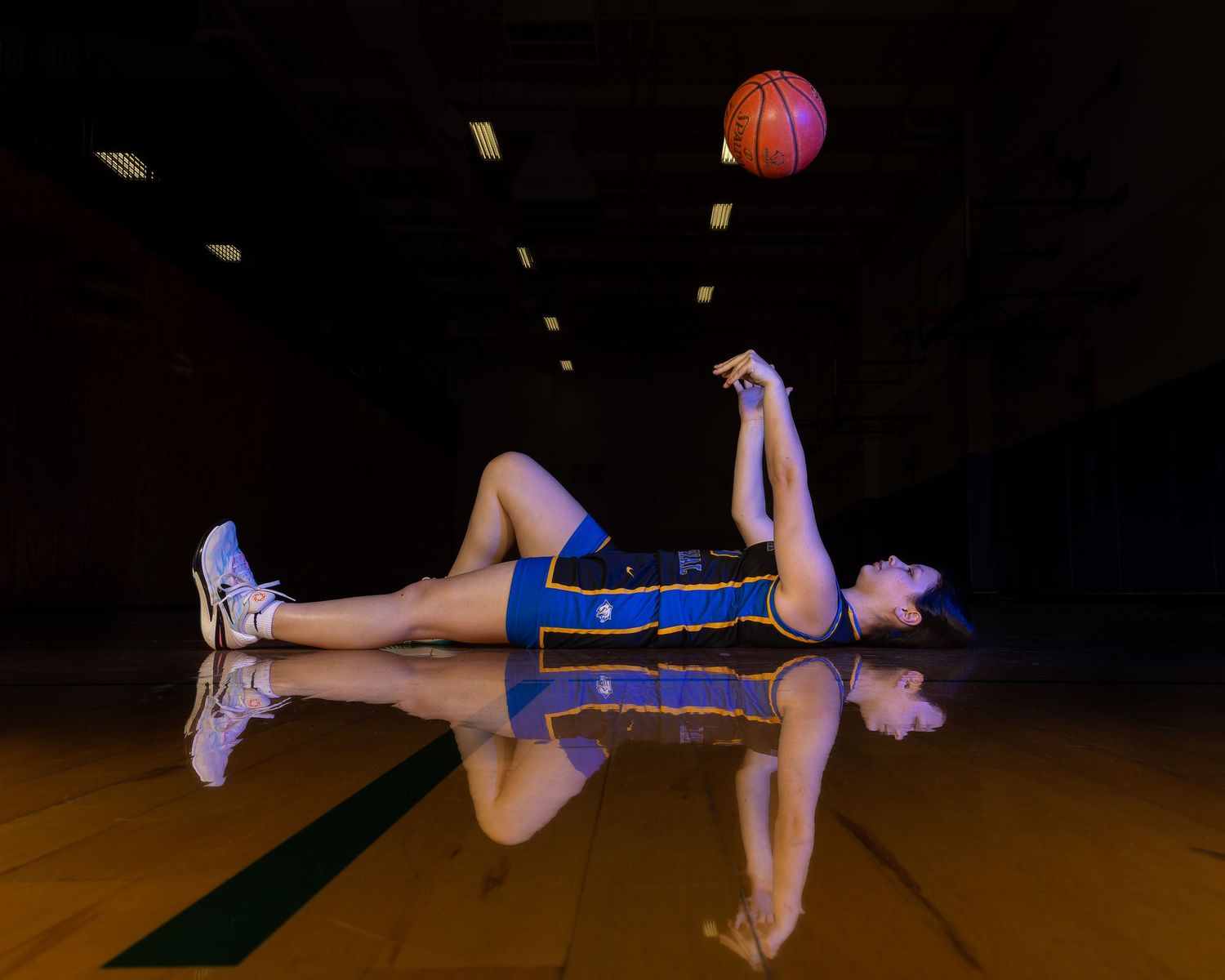 Basketball player lying on reflective court surface balancing ball above in blue uniform.