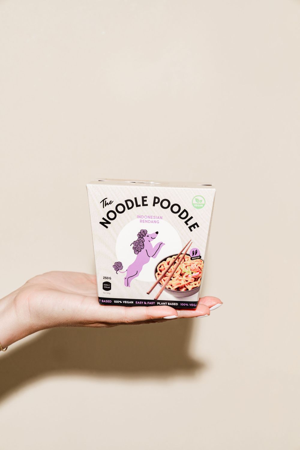 Hand holding a Noodle Poodle branded food package against a beige background.