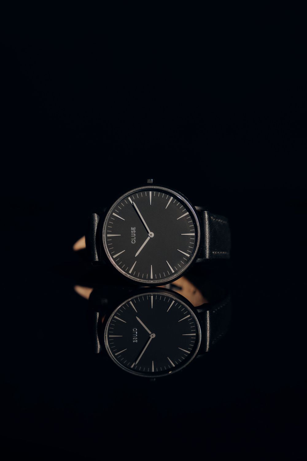 Two minimalist black watches with leather straps reflected on dark surface.