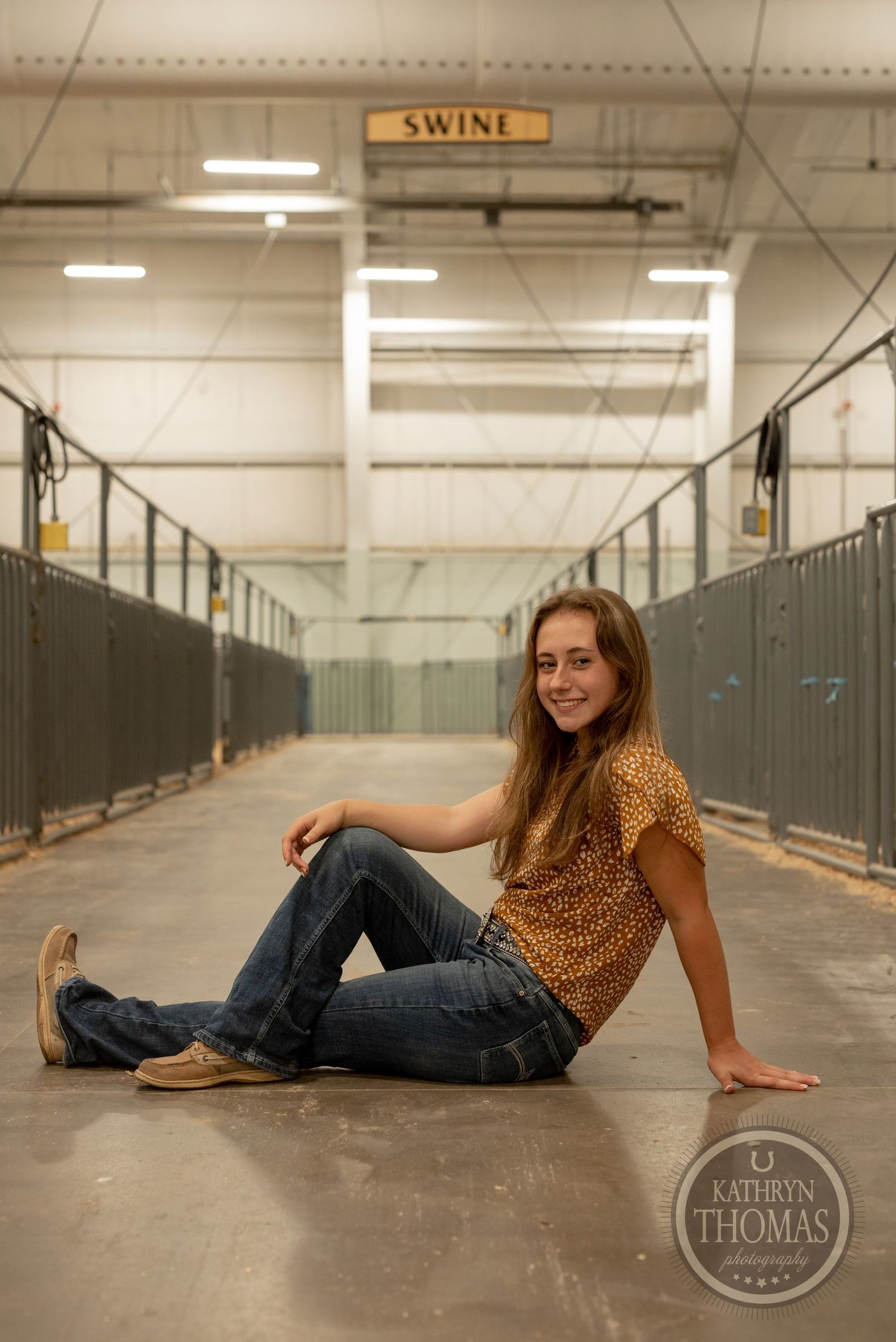 Casual sitting pose in livestock facility hallway wearing animal print top and jeans.