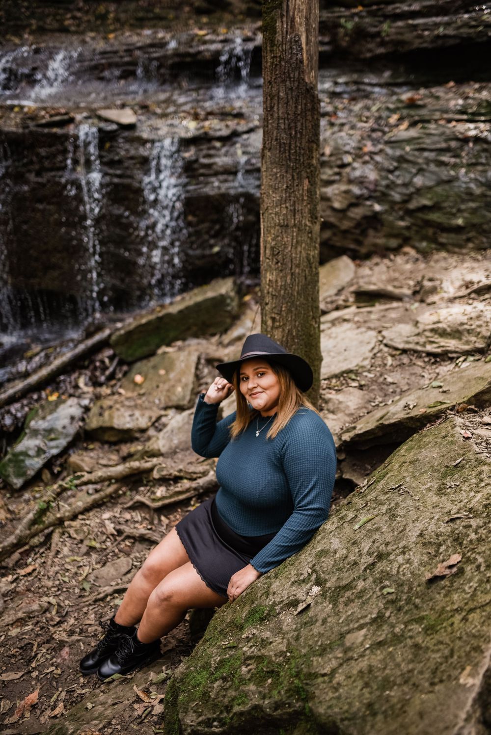 Figure in dark clothing reclines on mossy rocks by a waterfall.