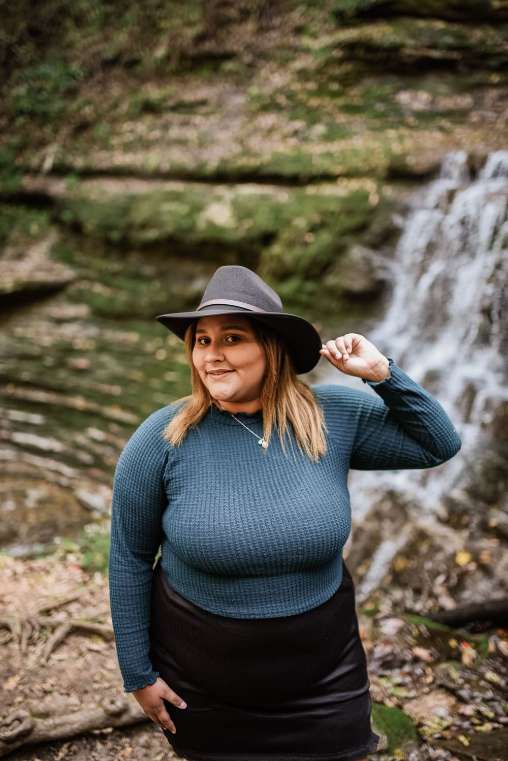 Person in blue sweater and black hat poses near cascading waterfall.