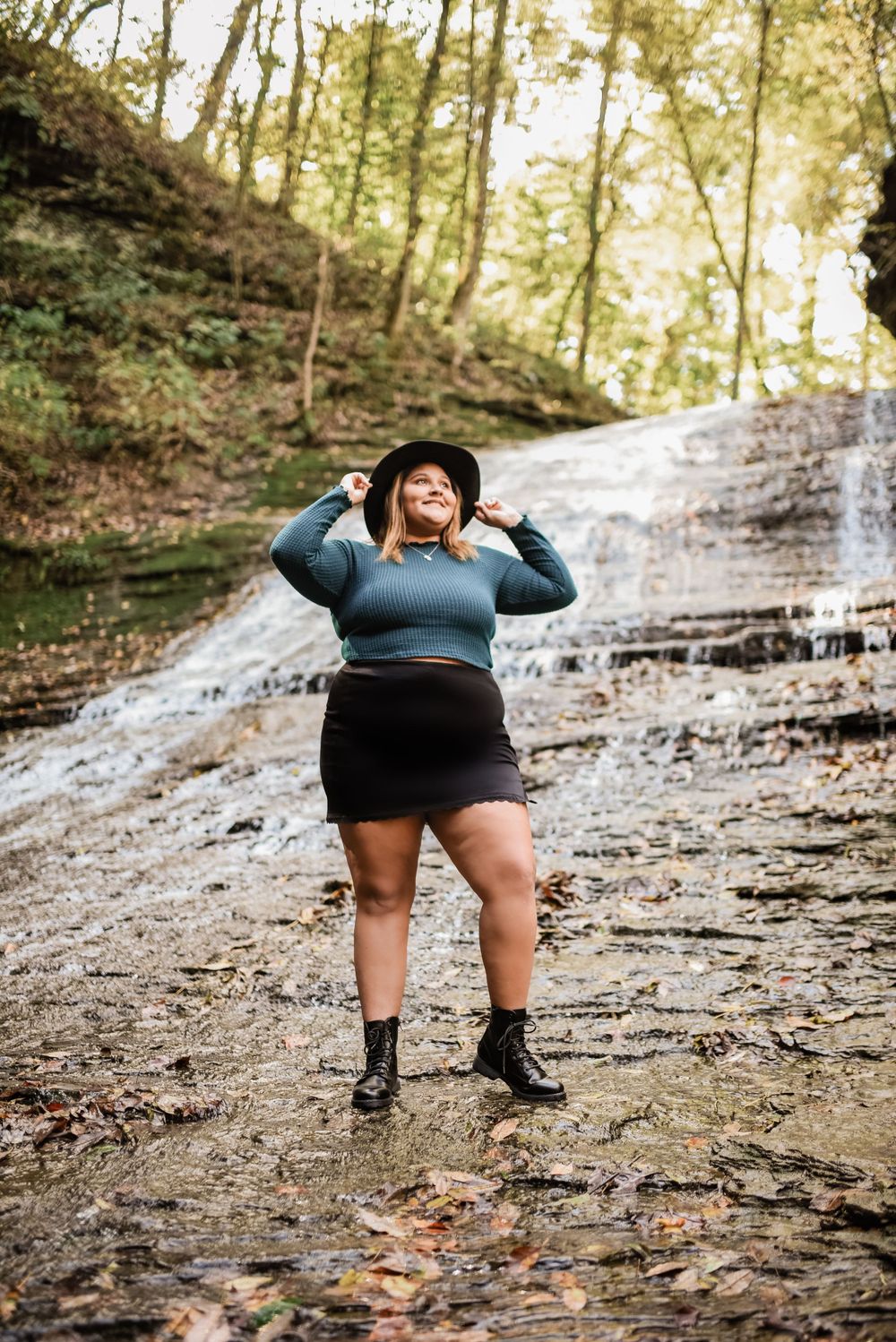Figure in dark outfit strikes playful pose near flowing waterfall.