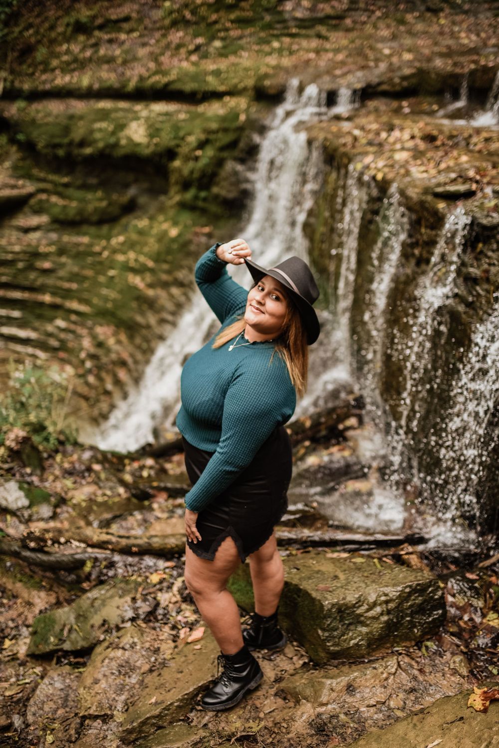 Figure in teal sweater poses confidently by cascading waterfall.