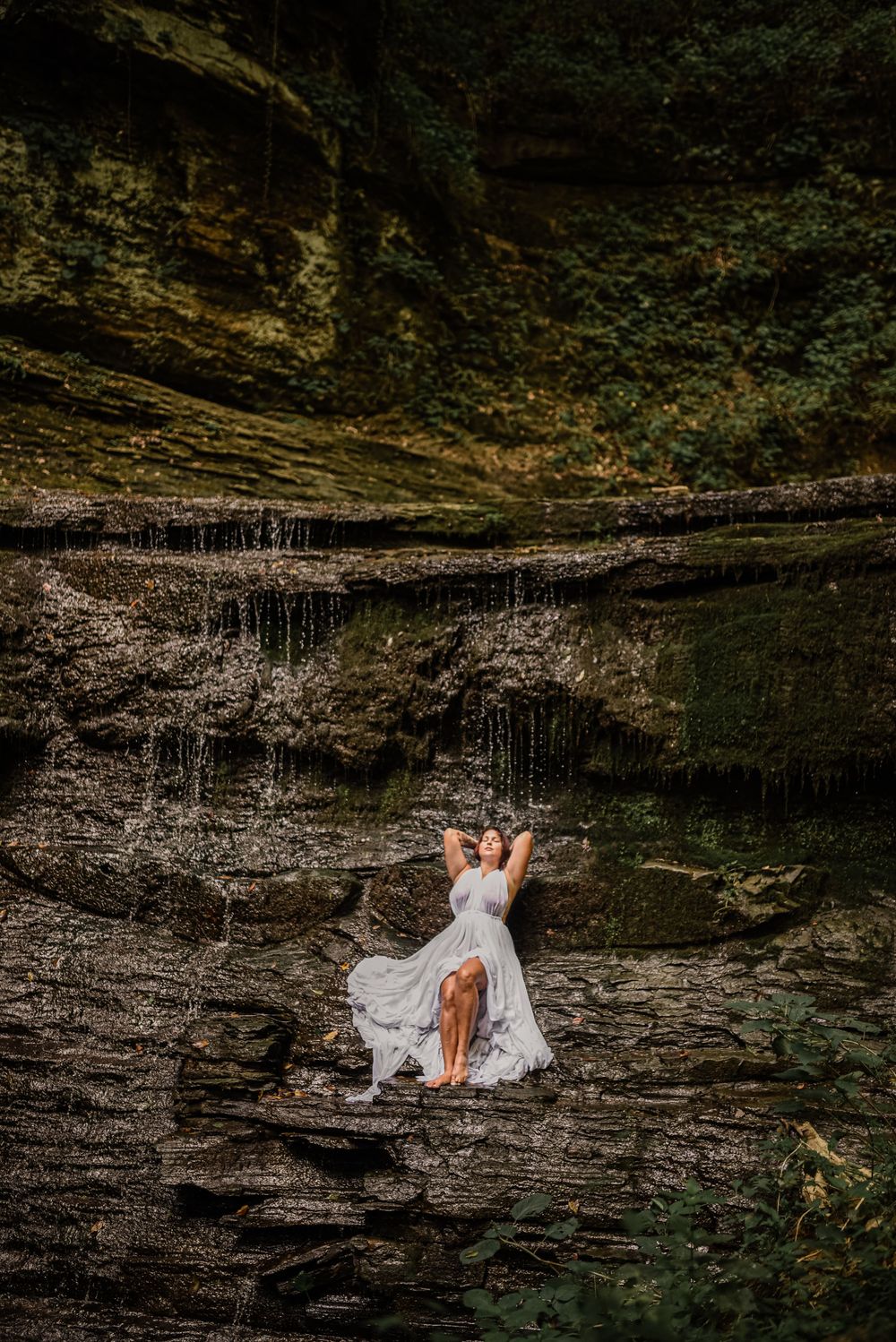 A dramatic waterfall cliff face serves as backdrop for an artistic portrait in flowing white fabric.